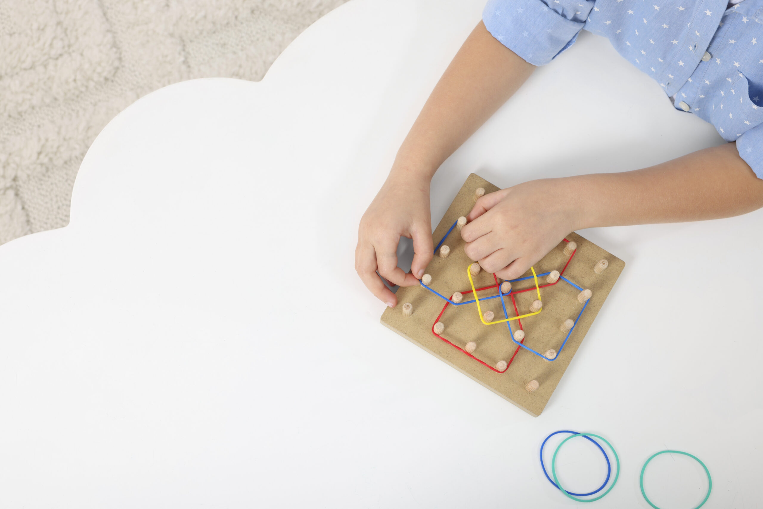 Motor skills development. Girl playing with geoboard and rubber bands at white table, top view. Space for text